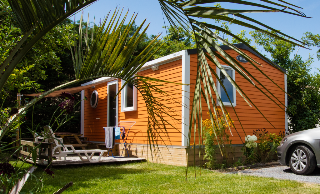 Mobil-home orange avec terrasse en bois, jardin verdoyant.